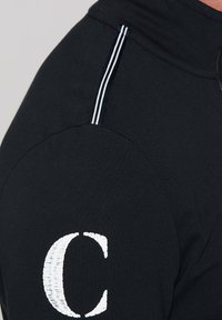 Sudadera negra con un alto cuello, rayas blancas en contraste en los hombros y una letra "C" bordada en blanco en la parte frontal.