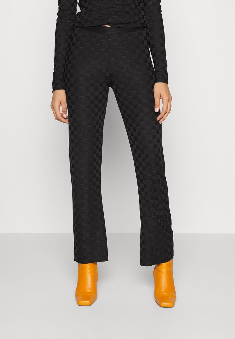 Femme portant un pantalon noir à carreaux taille haute et un haut à manches longues assorti, associé à des bottines orange vif, debout sur un sol gris.