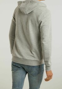 Hombre vistiendo una sudadera gris clara y jeans azules desgastados, mostrado de espaldas con el brazo derecho relajado a un lado.