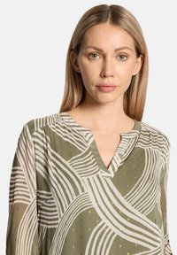 Grün-weiß gemusterte Bluse mit V-Ausschnitt, mit abstrakten wellenförmigen Streifen und goldenen Akzenten auf einem glatten Stoff.