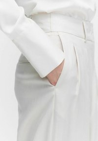 Witte op maat gemaakte broeken met gedetailleerde plooien, hoge tailleband en zijzakken, gemaakt van gladde, lichte stof. Lange witte blouse is zichtbaar.