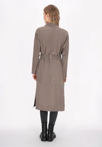 Slank, lang taupe trenchcoat med bælte i taljen, sideslidser og krave, fremstillet af glat stof. Båret over mørke leggings og støvler.