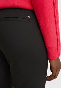 Högra handen på en person i svarta byxor med bakficka och en röd stickad tröja är synlig mot en enkel bakgrund.