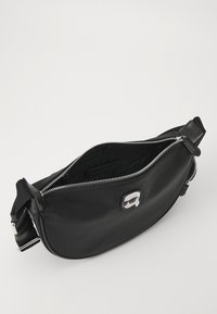 Černá crossbody taška se stříbrným zipem, nastavitelným popruhem a malým logem osoby nosící sluneční brýle na přední straně. Interiér je částečně viditelný.