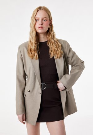 Frau mit langen, welligen blonden Haaren, trägt einen beigen Blazer über einem schwarzen, gerippten Kleid mit Gürtel, steht vor einem einfarbigen weißen Hintergrund.