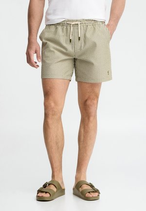 Mężczyzna ubrany w beżowo-białe krótkie spodenki w paski z wiązanym paskiem w talii, białą koszulę i oliwkowozielone sandały na klamrę, stojący na tle białego tła.