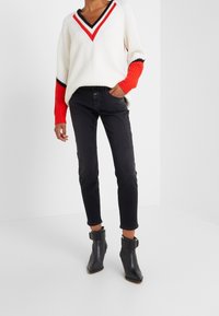 Persona vistiendo un suéter blanco de cuello en V con acentos rojos y negros, jeans ajustados negros y botas negras hasta el tobillo con hebillas.