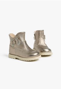 Metallisch goldene Knöchelboots mit einer strukturierten Oberfläche, einem dekorativen Bogen an der Seite und einer hell gefärbten Gummisohle.