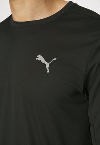 Svart texturerad skjorta som bärs av en person, med en liten silverfärgad Puma-logga på vänster bröstområde.