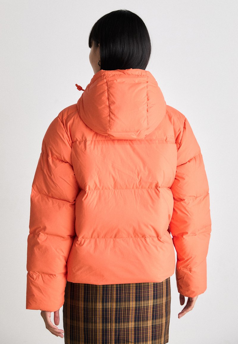 Aufgeblasene Jacke mit Kapuze in leuchtendem Orange, mit einem gesteppten Design, elastischen Bündchen und einer glatten Textur. Über einem karierten Rock getragen.