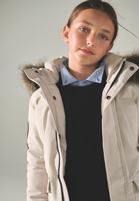 Khaki parka jaka ar pūkainu apdari uz kapuces, melna adīta džemperis zem tā un gaiši zila krekla ar apkakli. Gluda tekstūra, rāvējslēdzējs un pogu aizdare.