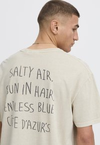 Beige oversized t-shirt featuring black text on the back: "SALTY AIR, SUN IN HAIR, ENDLESS BLUE, CÔTE D'AZUR'S." Soft, cotton material.