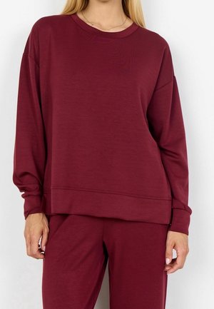 Kvinde iført en langærmet, løstsiddende vinrød sweatshirt og matchende bukser, stående mod en ensfarvet hvid baggrund.