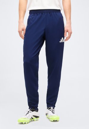 Mężczyzna ubrany w granatowe spodnie dresowe Adidas z białym logo oraz białe buty sportowe z neonowożółtymi korkami, stojący na tle jednolitym.