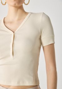 Haut beige à manches courtes côtelé avec un décolleté en V et trois boutons. Texture douce et design ajusté, longueur courte, ourlet soigneusement fini.