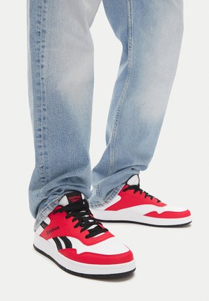 Osoba ubrana w jasnoniebieskie dżinsy oraz czerwono-białe-czarne buty Reebok z czarnymi sznurówkami, stojąca na białym tle.