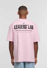 Roosa puuvillane t-särk lühikeste varrukatega, mille seljal on silmapaistev must grafikatekst "LEADERS' LAB." Lihtne disain.