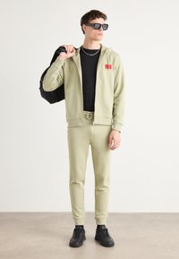 Tuta verde pallido con felpa con zip e pantaloni con cordino, abbinata a una maglietta nera e sneaker nere. Borsa nera e occhiali da sole.