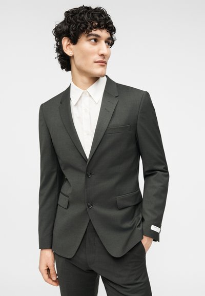 Σκούρο γκρι κοστούμι με δομημένη εφαρμογή, διαθέτει πέτο με εγκοπές, δύο κουμπιά και τσέπες. Συνδυάζεται με λευκό πουκάμισο με κουμπιά.