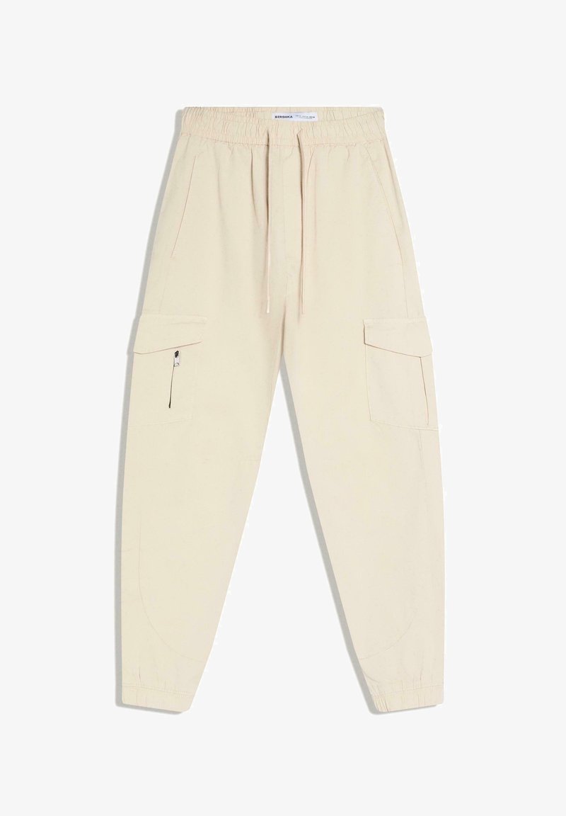 Pantalon cargo jogger beige avec taille élastique, cordon de serrage, poches à rabat latérales, poche zippée sur la jambe gauche et poignets élastiques aux chevilles.