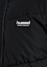 Svart vadderad jacka med en kviltad textur, smidig dragkedja och en framträdande vit logotypmärke med texten "hummel SPORTSWEAR."