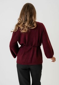 Pulover tricotat bordeaux cu talie legată, mâneci lungi tip balon, țesătură texturată și manșete canalate, asortat cu pantaloni negri.