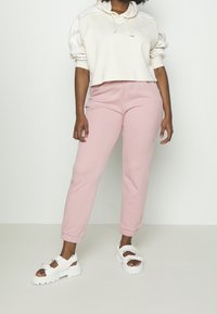 Krátká krémová mikina s kapucí a šňůrkami, bílé pruhy; růžové tepláky s elastickými manžetami; bílé sandály s perlovými ozdobami a tlustou podrážkou.