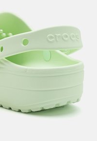 Grön gummiklump med tjock sula, justerbar rem och cirkulära ventilationshål. Logotypen "crocs" är präglad på remmen.