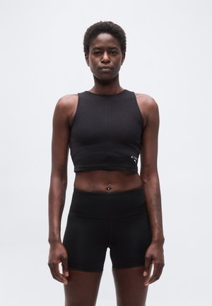 Velformet person med kort, krøllet hår står vendt fremad iført en sort ærmeløs crop-top og sorte shorts med høj talje mod en ensfarvet baggrund.