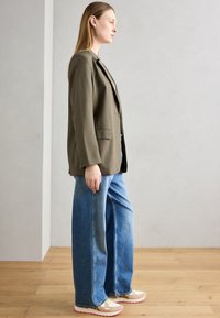 Blazer sartorial verde oliva, camicia grigia chiara, jeans blu a vestibilità rilassata e scarpe beige con suole rosa e dettagli decorativi.