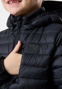 Sort polstret jakke med høj krave, lynlås-lukning og logo-patch. Har en glat tekstur og elastiske manchetter. Let quiltet design.