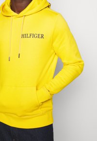 Felpa gialla con cappuccio con coulisse e logo "HILFIGER" in blu navy. Tessuto liscio, tasca kangaroo, polsini a costine, dettaglio accentuato vicino all'orlo.
