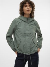 Frau trägt eine salbeigrüne Kapuzenjacke mit Reißverschluss, Druckknopftaschen, elastischen Bündchen und Tunnelzug am Saum, steht vor einem weißen Hintergrund.