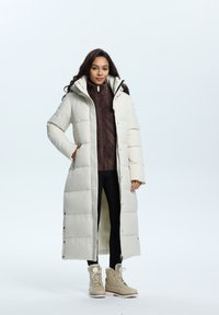 Lange crèmekleurige pufferjas met capuchon, voorzien van een ritssluiting aan de voorkant, zwarte broek en tan-kleurige winterlaarzen. Daaronder is een donkerbruin gebreid sweater zichtbaar.