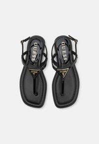 Zwarte platte sandalen met een glanzende afwerking, voorzien van kruislings gespen en een driehoekig metalen logo-accent op de teenband.