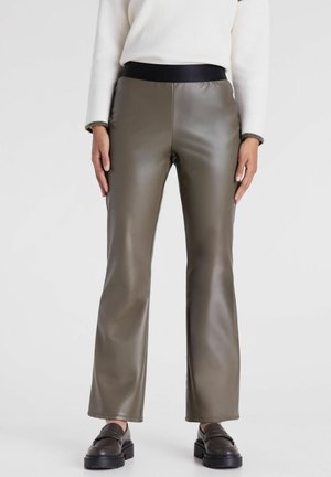 Vrouw draagt olijfgroene nep leren broek met zwarte elastische tailleband en donkerbruine chunky loafers, staand tegen een witte achtergrond.
