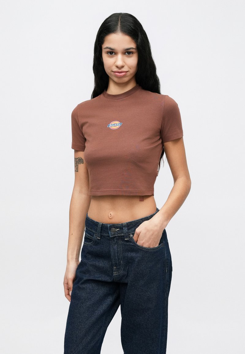 Femme aux longs cheveux noirs portant un t-shirt marron court avec un logo et un jean bleu foncé ample, posant avec une main dans la poche.
