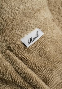 Beiges Handtuch mit einer strukturierten, flauschigen Oberfläche. Es hat ein genähtes weißes Etikett mit "Reell" in schwarzer Kursive. Weicher und saugfähiger Stoff.