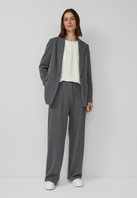 Frau trägt einen grauen Nadelstreifenanzug mit passenden weit geschnittenen Hosen und Blazer, weiße Bluse und weiße Sneakers, steht vor einfachem Hintergrund.