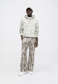 Világos bézs kapucnis pulóver finom mintázattal, bézs árnyalatú, bőszárú mintás nadrággal párosítva. A fehér sportcipők egészítik ki a megjelenést.