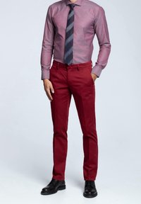 Červené bavlněné kalhoty s úzkým střihem, spojené s texturovanou světle fialovou košilí a pruhovanou tmavou kravatou. Černé šněrovací boty dotvářejí vzhled.