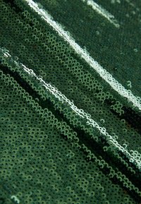 Gros plan d’un tissu vert densément couvert de petits sequins brillants reflétant la lumière en motifs diagonaux.