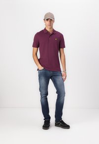 Burgundská polo košeľa so žltým logom, v kombinácii s modrými džínami. Čierne tenisky dotvárajú vzhľad. Model stojí na bielom pozadí.