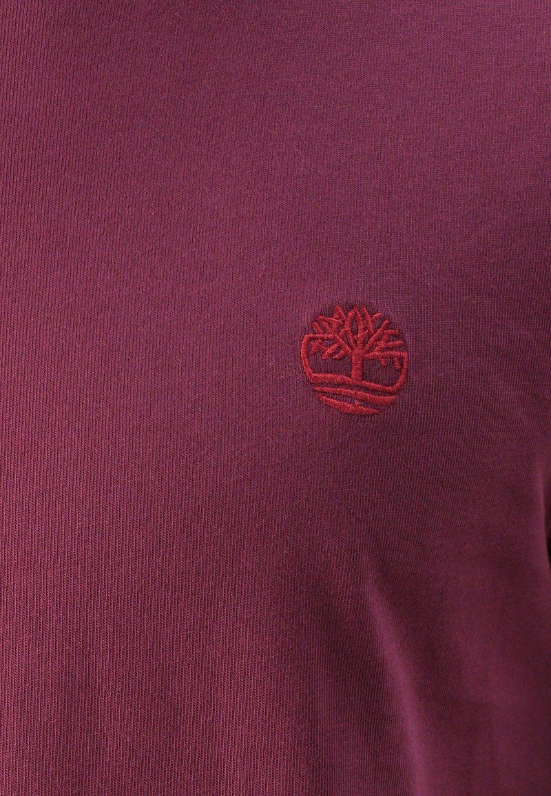 Maglione a collo alto color bordeaux, con texture a costine e un logo rosso ricamato a forma di albero sul lato sinistro del petto.