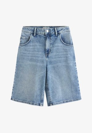 Dziewczyna stoi, ubrana w żółtą bluzkę w kwiaty z krótkim rękawem, krótkie spodenki dżinsowe do kolan i białe trampki na jednolitym tle.