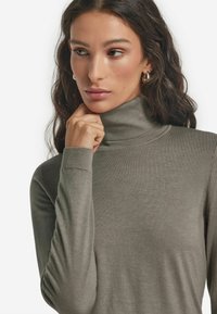 Grå turtlenecktröja i slät tyg, med lång ärmar och ribbade muddar. Modellens hand vilar på hakan och visar halsringningen.