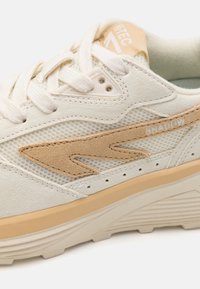 White sneaker with a mesh upper, suede beige accents, white laces, and a textured tan outsole. Features "SHADOW" branding in white.
