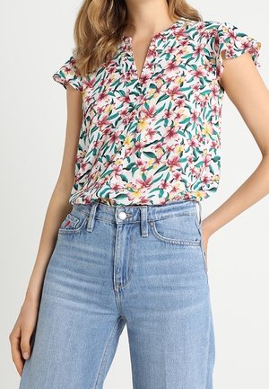 Woman wearing a short-sleeve floral blouse with pink and yellow flowers paired with high-waisted light blue jeans, hand resting on hip.