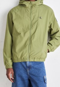 Lätt olivgrön jacka med huva, elastiska ärmmuddar och nederkant, samt en logotyp på bröstet. Bärs över blåa cargobyxor.