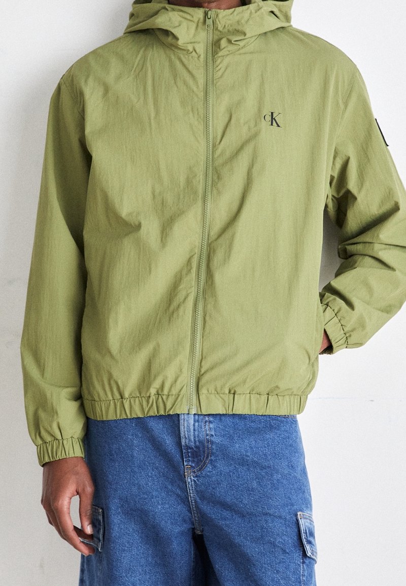 Lätt olivgrön jacka med huva, elastiska ärmmuddar och nederkant, samt en logotyp på bröstet. Bärs över blåa cargobyxor.